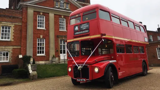 Red vintage Routemaster wedding bus decorated with white ribbons parked outside a historic red brick venue.