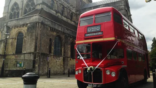 Red Routemaster double decker bus decorated with white wedding ribbons parked outside a historic church.