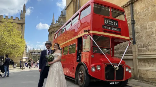 Bride and groom standing beside a red Routemaster double decker wedding bus with white ribbons outside a cathedral.