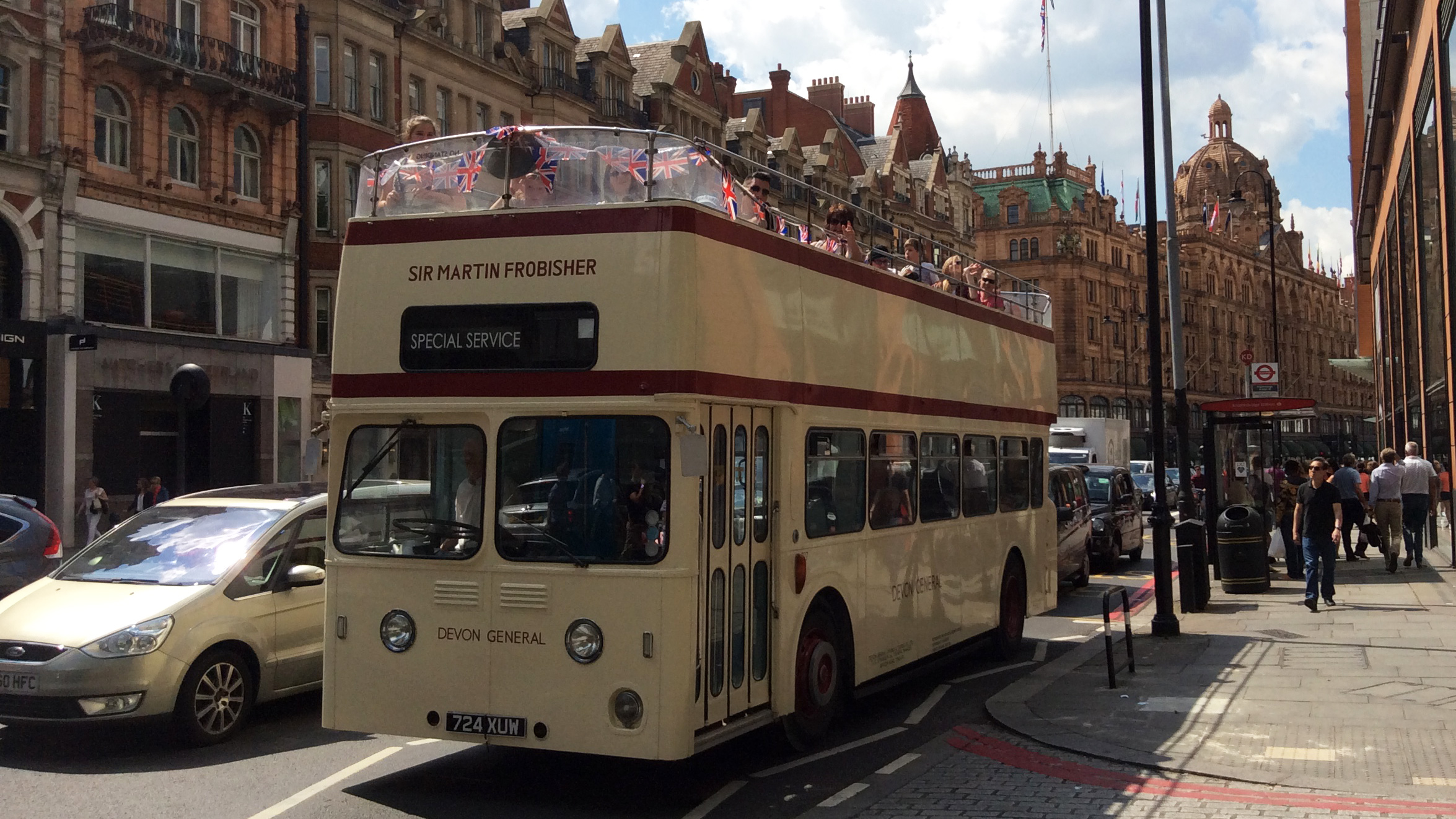 Cream vintage Leyland double decker wedding bus driving through a city street with historic buildings.