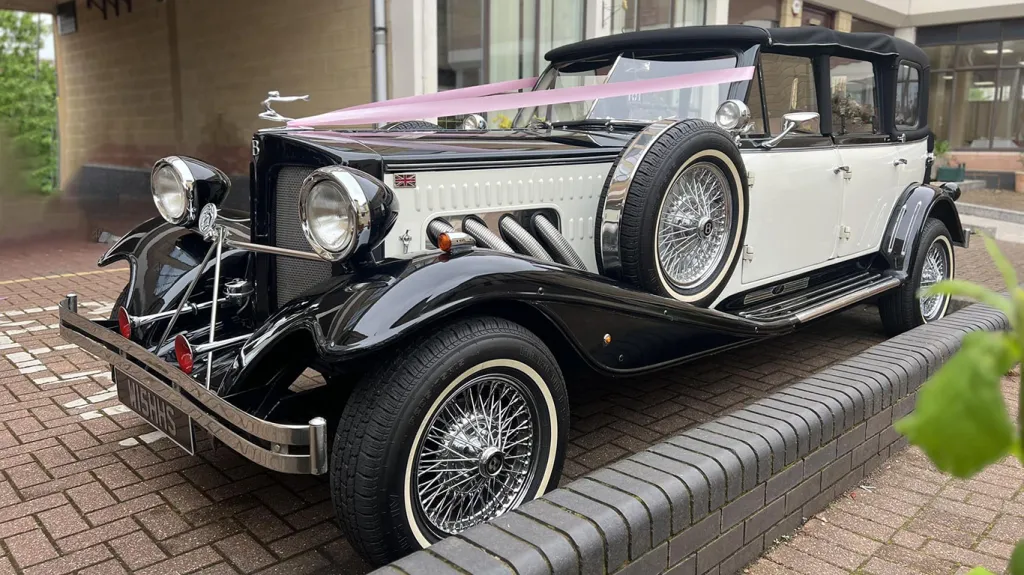 Black and white vintage Beauford wedding car with chrome grille, chrome headlamps, pink ribbons and white wall tyres parked outside a London venue entrance on a brick driveway.