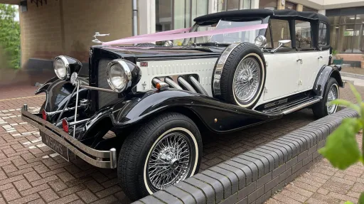 Black and white vintage Beauford wedding car with chrome grille, chrome headlamps, pink ribbons and white wall tyres parked outside a London venue entrance on a brick driveway.