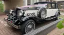 Black and white vintage Beauford wedding car with chrome grille, chrome headlamps, pink ribbons and white wall tyres parked outside a London venue entrance on a brick driveway.
