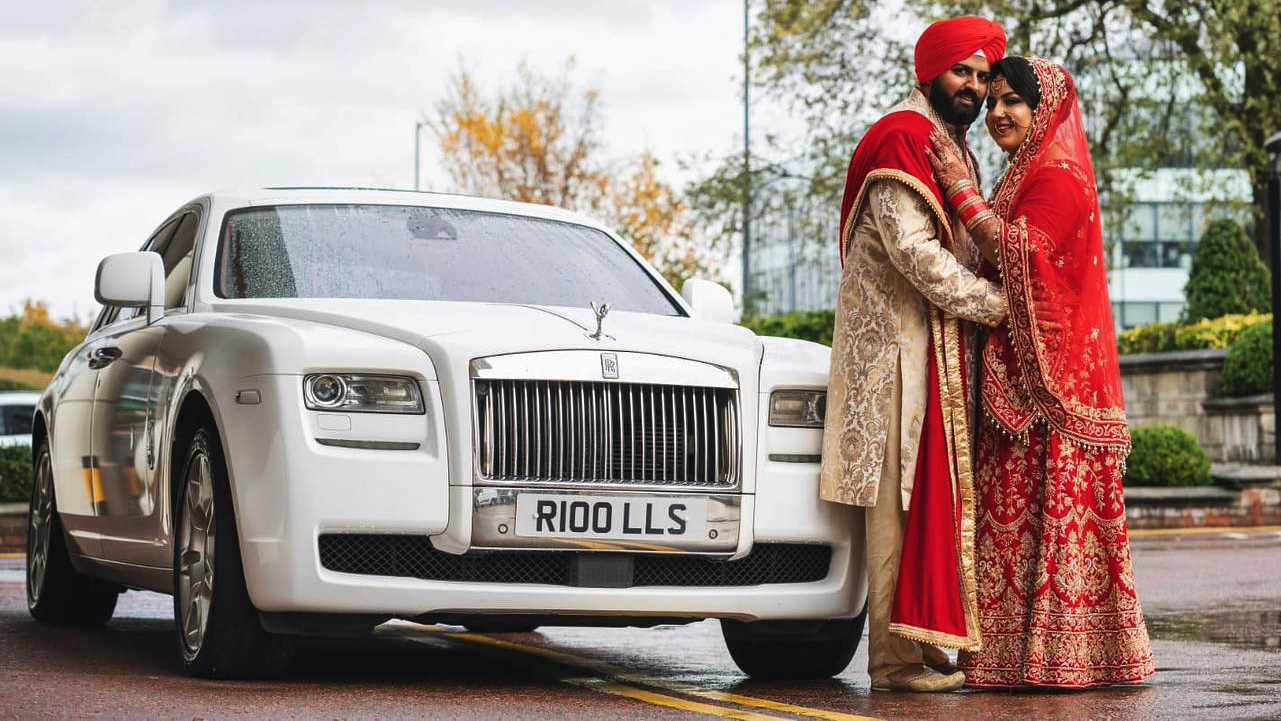 Asian bride and groom standing beside a white Rolls-Royce Ghost luxury wedding car with polished chrome grille, Spirit of Ecstasy bonnet ornament and chrome alloy wheels parked on a London street.