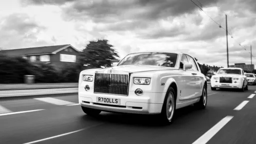 Black and white photo of a white Rolls-Royce Phantom luxury wedding car driving along a city road with chrome grille, Spirit of Ecstasy bonnet ornament and polished alloy wheels.