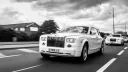 Black and white photo of a white Rolls-Royce Phantom luxury wedding car driving along a city road with chrome grille, Spirit of Ecstasy bonnet ornament and polished alloy wheels.