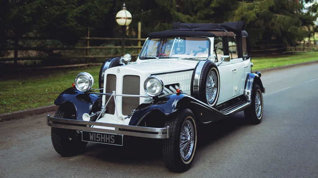 Black and ivory vintage Beauford tourer wedding car driving along a tree-lined country road, featuring chrome radiator grille, chrome wire wheels and classic white wall tyres