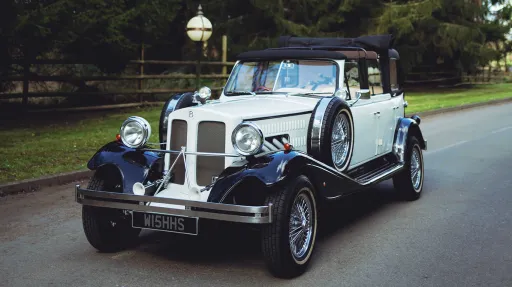 Black and ivory vintage Beauford tourer wedding car driving along a tree-lined country road, featuring chrome radiator grille, chrome wire wheels and classic white wall tyres