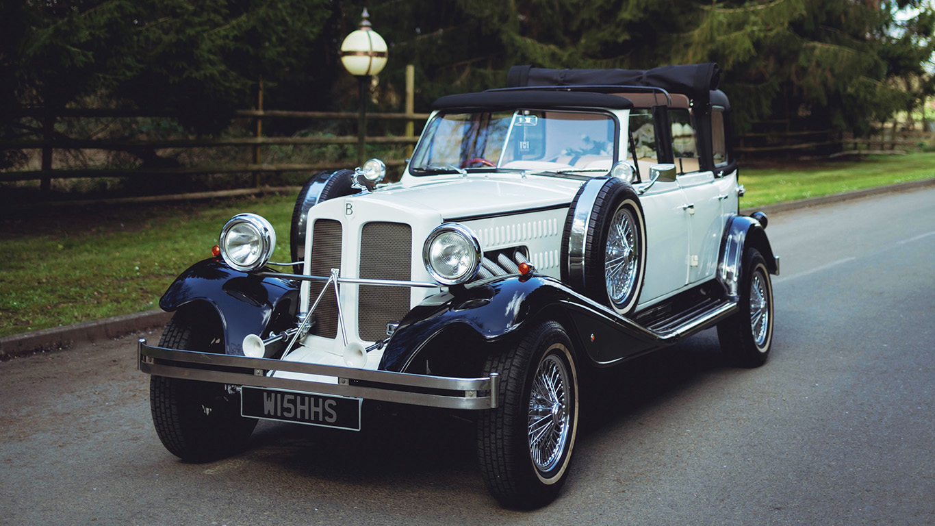Black and ivory vintage Beauford tourer wedding car driving along a tree-lined country road, featuring chrome radiator grille, chrome wire wheels and classic white wall tyres