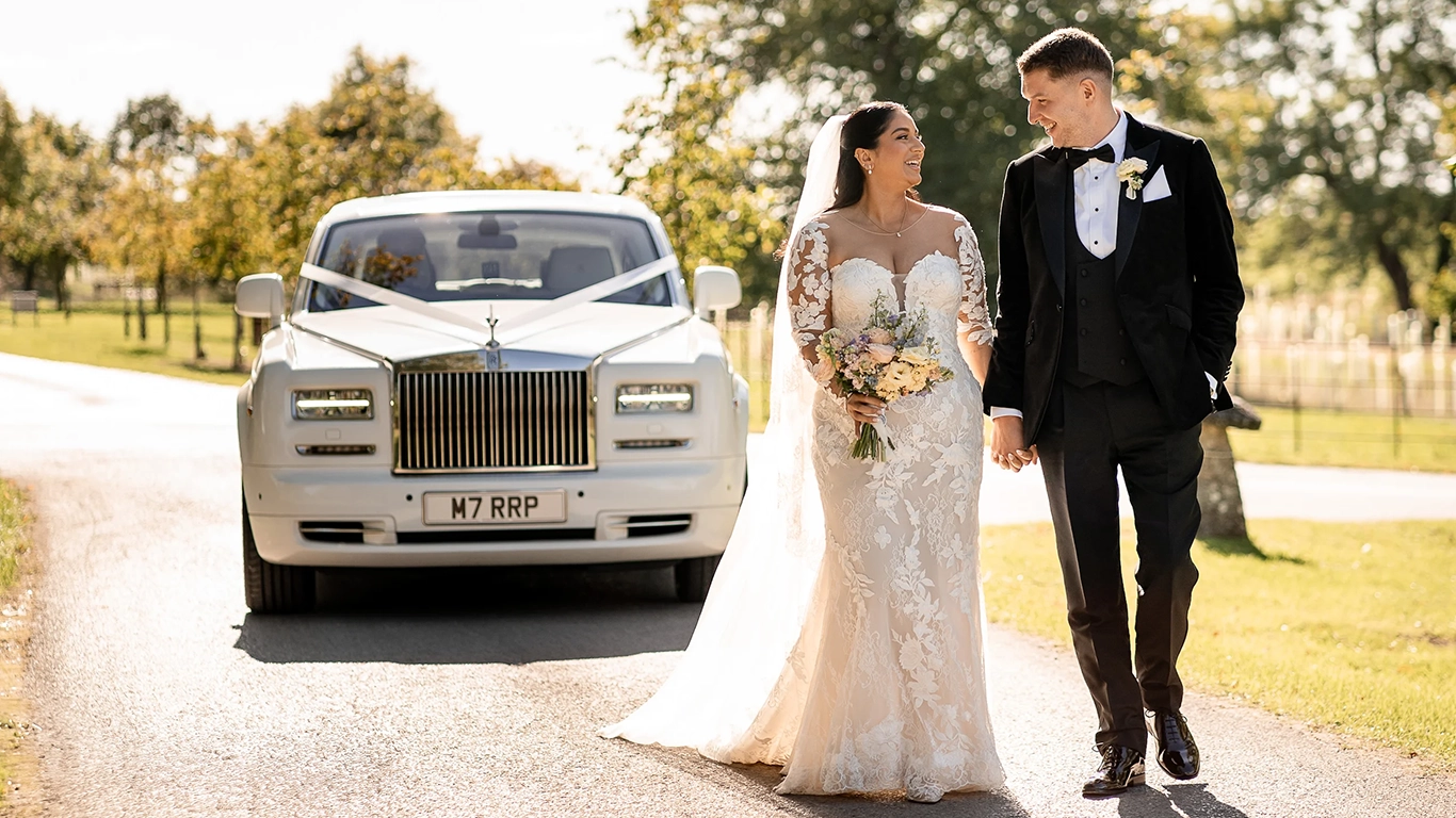 Bride and groom walking in front of a white Rolls-Royce Phantom Series II wedding car decorated with wedding ribbons, featuring chrome grille, Spirit of Ecstasy bonnet ornament and alloy wheels on a country road.