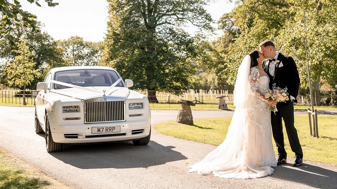 Bride and groom kissing beside a white Rolls-Royce Phantom Series II luxury wedding car with chrome grille, Spirit of Ecstasy bonnet ornament and polished alloy wheels parked beside trees.