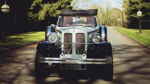Front view of a black and white Beauford vintage wedding car with polished chrome radiator, chrome headlights and white wall tyres parked on a shaded country lan