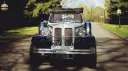 Front view of a black and white Beauford vintage wedding car with polished chrome radiator, chrome headlights and white wall tyres parked on a shaded country lan