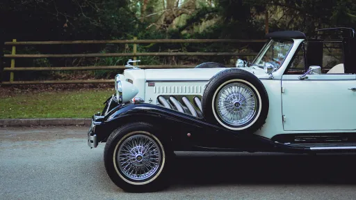 Side profile of a black and ivory Beauford vintage car showing long bonnet detail, chrome trim, chrome wire wheels and white wall tyres beside a rural roadside fence.