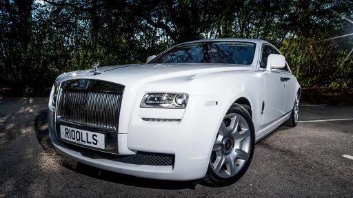 Front view of a white Rolls-Royce Ghost luxury chauffeur driven wedding car with chrome grille, Spirit of Ecstasy bonnet ornament and large alloy wheels parked on a driveway.