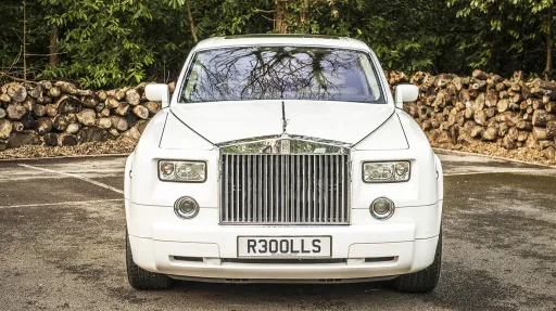 Full front view of a white Rolls-Royce Phantom luxury wedding car with polished chrome grille and alloy wheels parked on a driveway with stacked logs in the background.