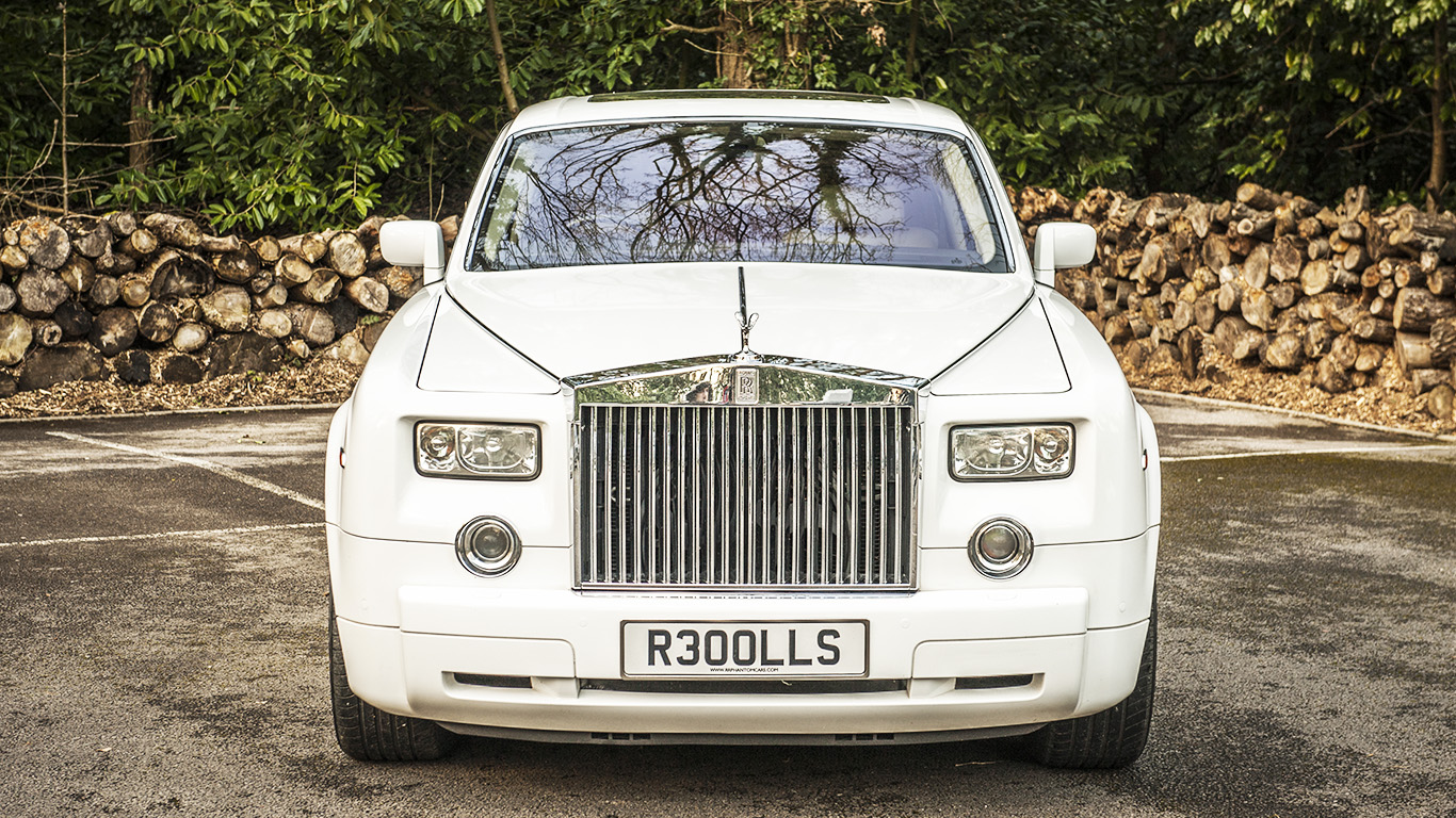 Full front view of a white Rolls-Royce Phantom luxury wedding car with polished chrome grille and alloy wheels parked on a driveway with stacked logs in the background.