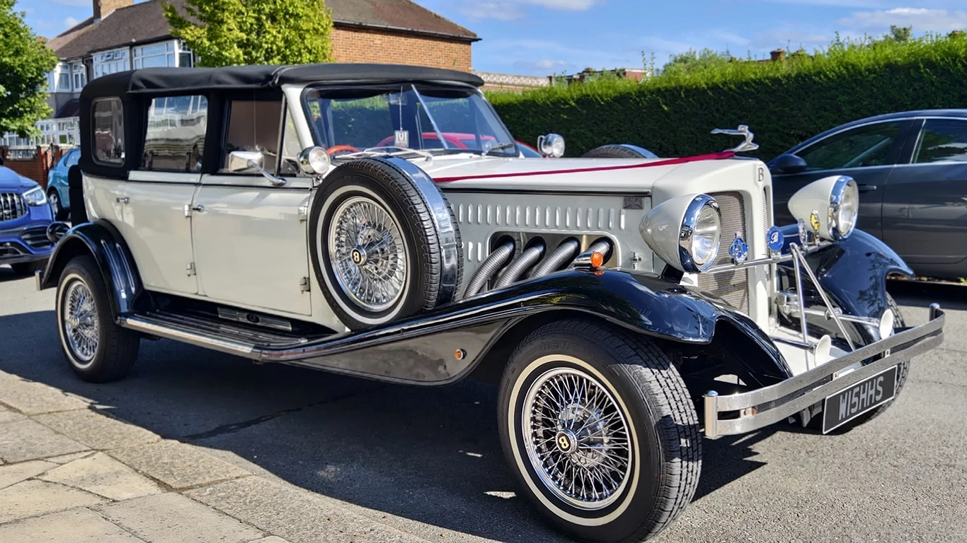 Full view of a black and white vintage Beauford semi-convertible tourer with roll-back soft roof, chrome grille, chrome wire wheels and white wall tyres parked outside a building.