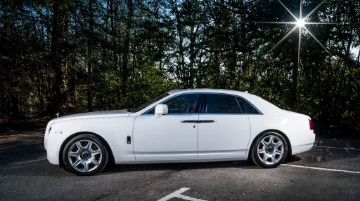 Side view of a white Rolls-Royce Ghost luxury wedding car with privacy rear windows, chrome window trim and polished alloy wheels parked beside a wooded road.