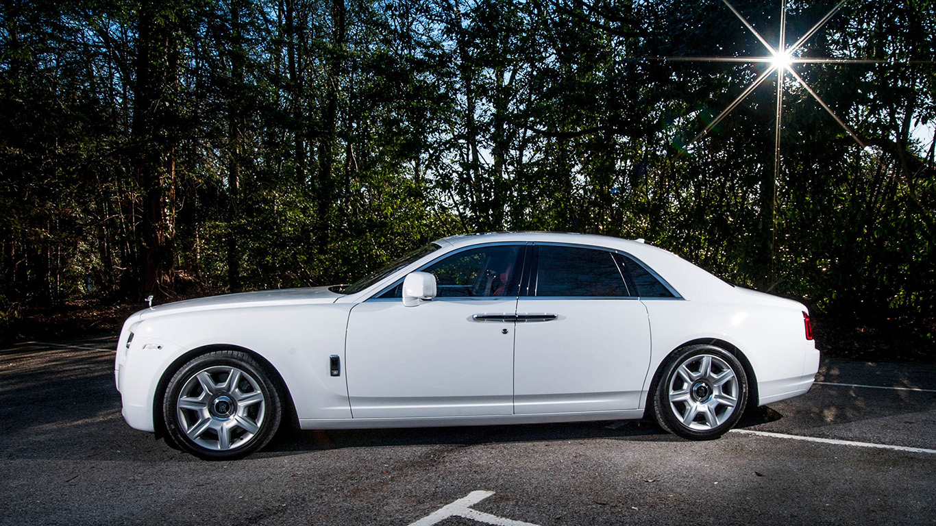 Side view of a white Rolls-Royce Ghost luxury wedding car with privacy rear windows, chrome window trim and polished alloy wheels parked beside a wooded road.