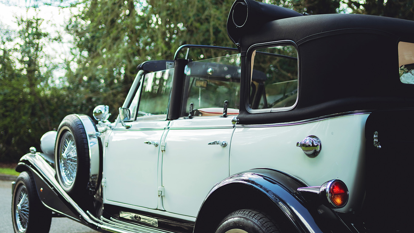 Side profile of a black and ivory Beauford vintage car showing long bonnet detail, chrome trim, chrome wire Full view of a black and white vintage Beauford semi-convertible tourer with roll-back soft roof, chrome grille, chrome wire wheels and white wall tyres parked outside a building.