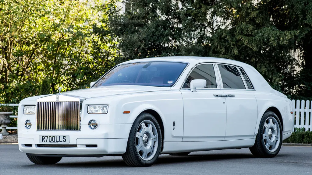 White Rolls-Royce Phantom luxury wedding car with polished chrome grille, Spirit of Ecstasy bonnet ornament and alloy wheels parked beside trees and greenery.