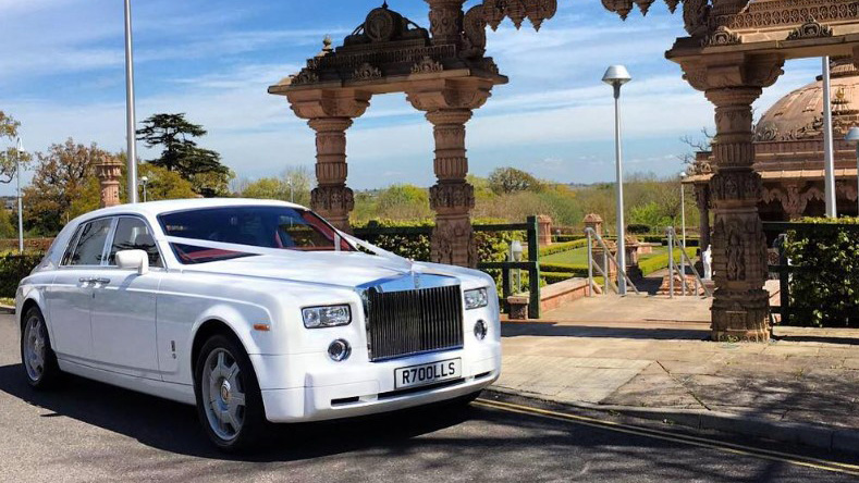 White Rolls-Royce Phantom luxury wedding car with chrome grille and polished alloy wheels parked outside an Asian wedding venue with decorative stone arches.
