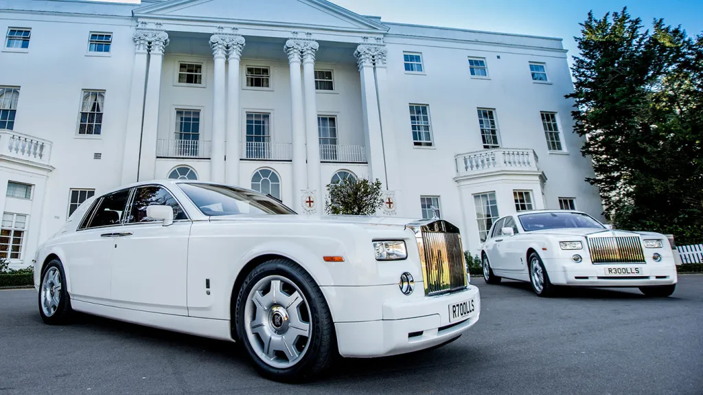 Two identical white Rolls-Royce Phantom luxury wedding cars with chrome grilles and alloy wheels parked outside a grand white mansion venue.