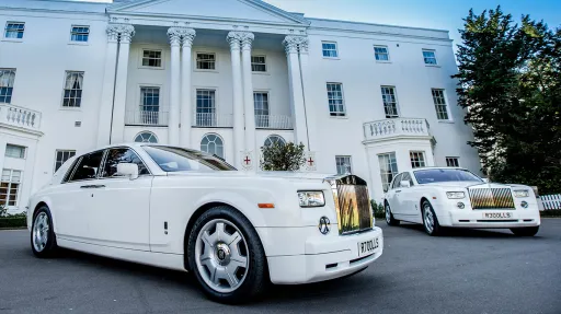 Two identical white Rolls-Royce Phantom luxury wedding cars with chrome grilles and alloy wheels parked outside a grand white mansion venue.