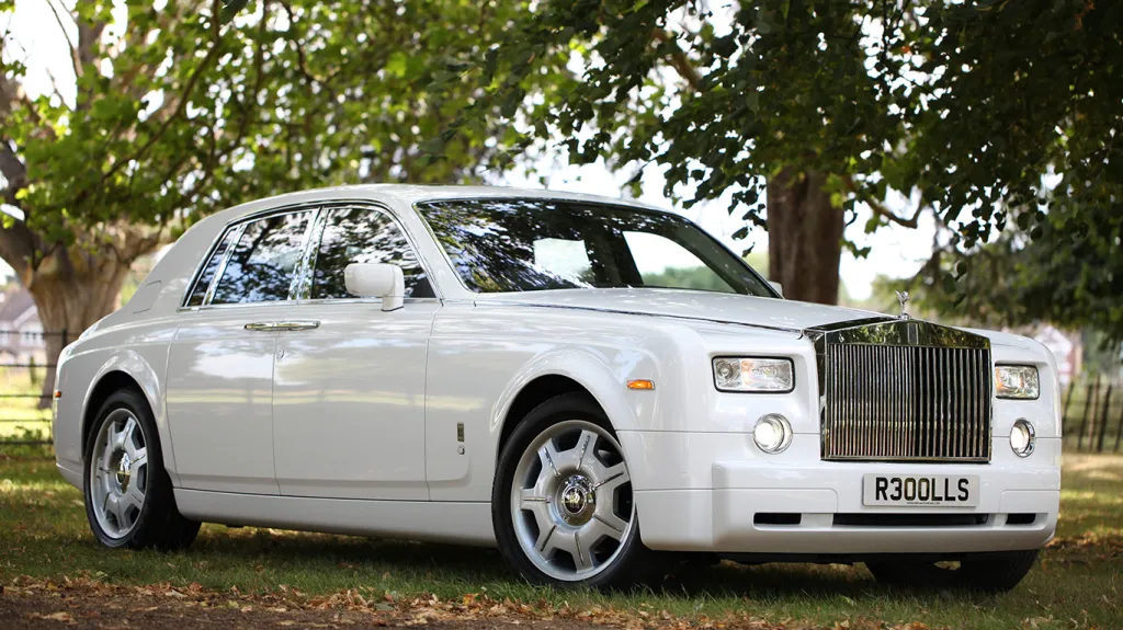 Side view of a white Rolls-Royce Phantom luxury chauffeur driven wedding car with chrome grille and alloy wheels parked beside trees and greenery.