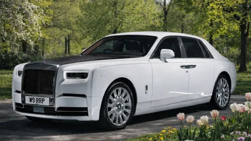 White Rolls-Royce Phantom VIII wedding car with grey bonnet, polished chrome grille, Spirit of Ecstasy bonnet ornament and large alloy wheels parked beside gardens and trees.v