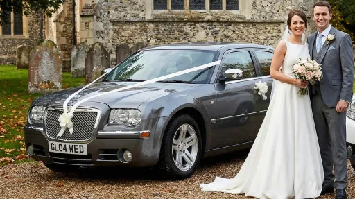 Newlywed couple posing beside a grey Chrysler 300C wedding car with white ribbon on the bonnet, parked outside a stone church on a gravel forecourt.