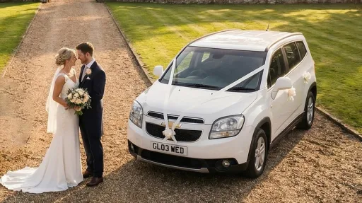 Bride and groom standing beside a white Chevrolet Orlando wedding car decorated with white ribbons, parked on a gravel driveway with trees and greenery in the background.