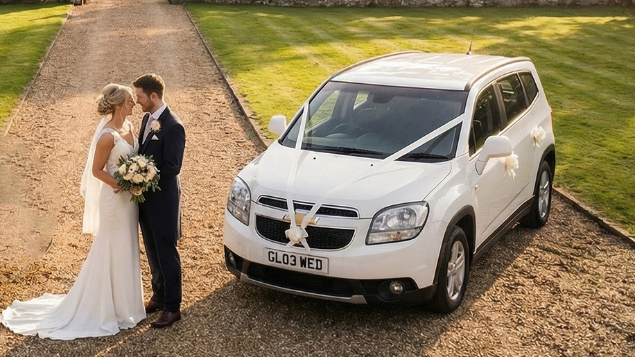 Bride and groom standing beside a white Chevrolet Orlando wedding car decorated with white ribbons, parked on a gravel driveway with trees and greenery in the background.