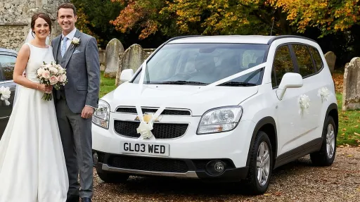 Newlywed couple posing beside a white Chevrolet Orlando wedding car with white ribbon on the bonnet, parked on a countryside driveway with autumn trees behind.