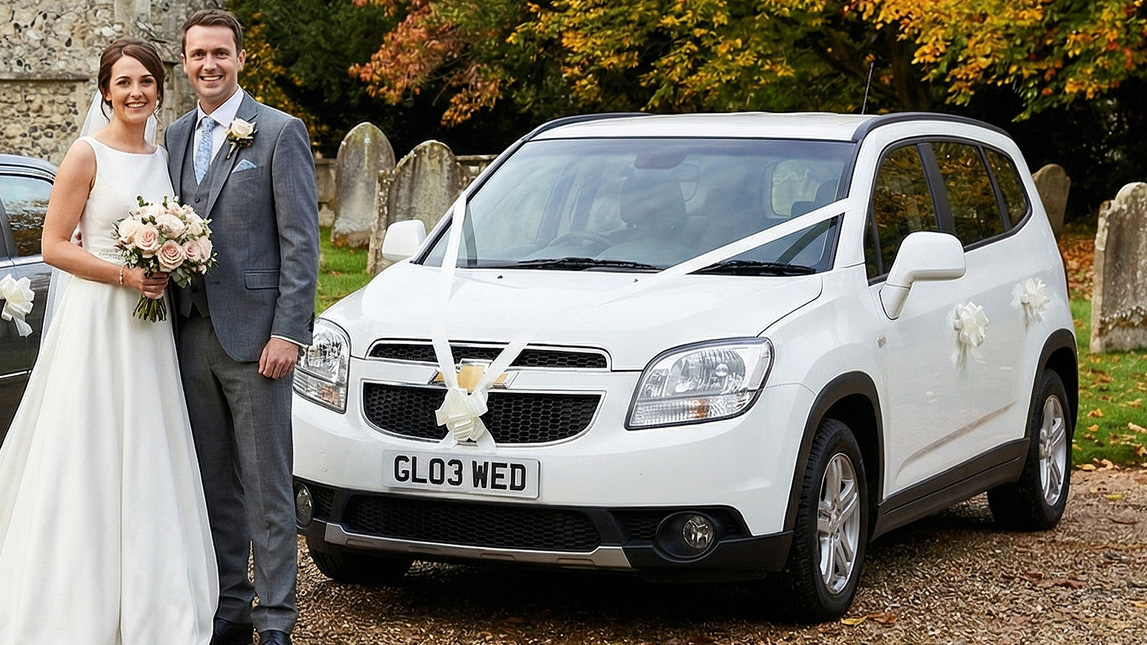 Newlywed couple posing beside a white Chevrolet Orlando wedding car with white ribbon on the bonnet, parked on a countryside driveway with autumn trees behind.