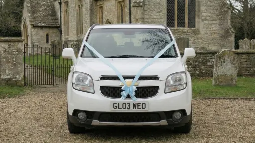 Front view of a white Chevrolet Orlando wedding car decorated with white ribbon across the bonnet, parked outside a stone church with gravel forecourt.