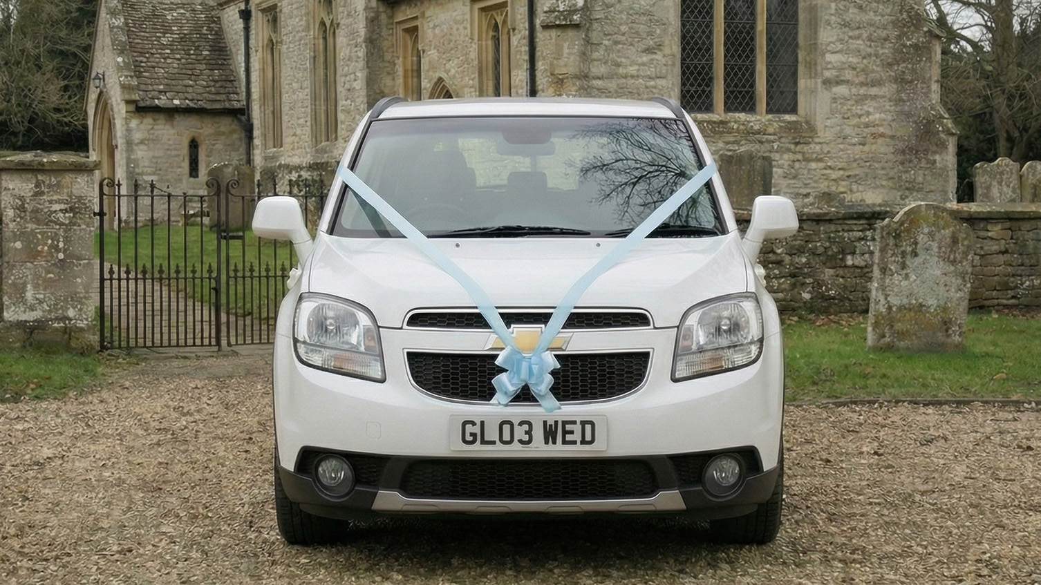 Front view of a white Chevrolet Orlando wedding car decorated with white ribbon across the bonnet, parked outside a stone church with gravel forecourt.
