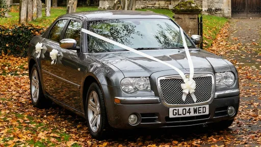 Grey Chrysler 300C wedding car with white ribbons across the bonnet parked on a leaf covered driveway with autumn trees and church buildings in the background.