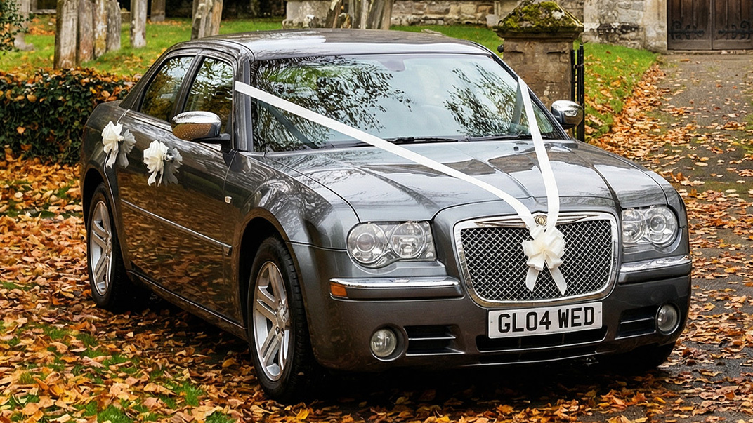 Grey Chrysler 300C wedding car with white ribbons across the bonnet parked on a leaf covered driveway with autumn trees and church buildings in the background.