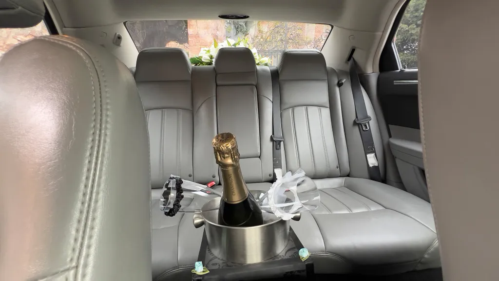 Rear interior of a Chrysler 300C wedding car showing light silver leather seats with champagne bottle and glasses placed between the rear seats.