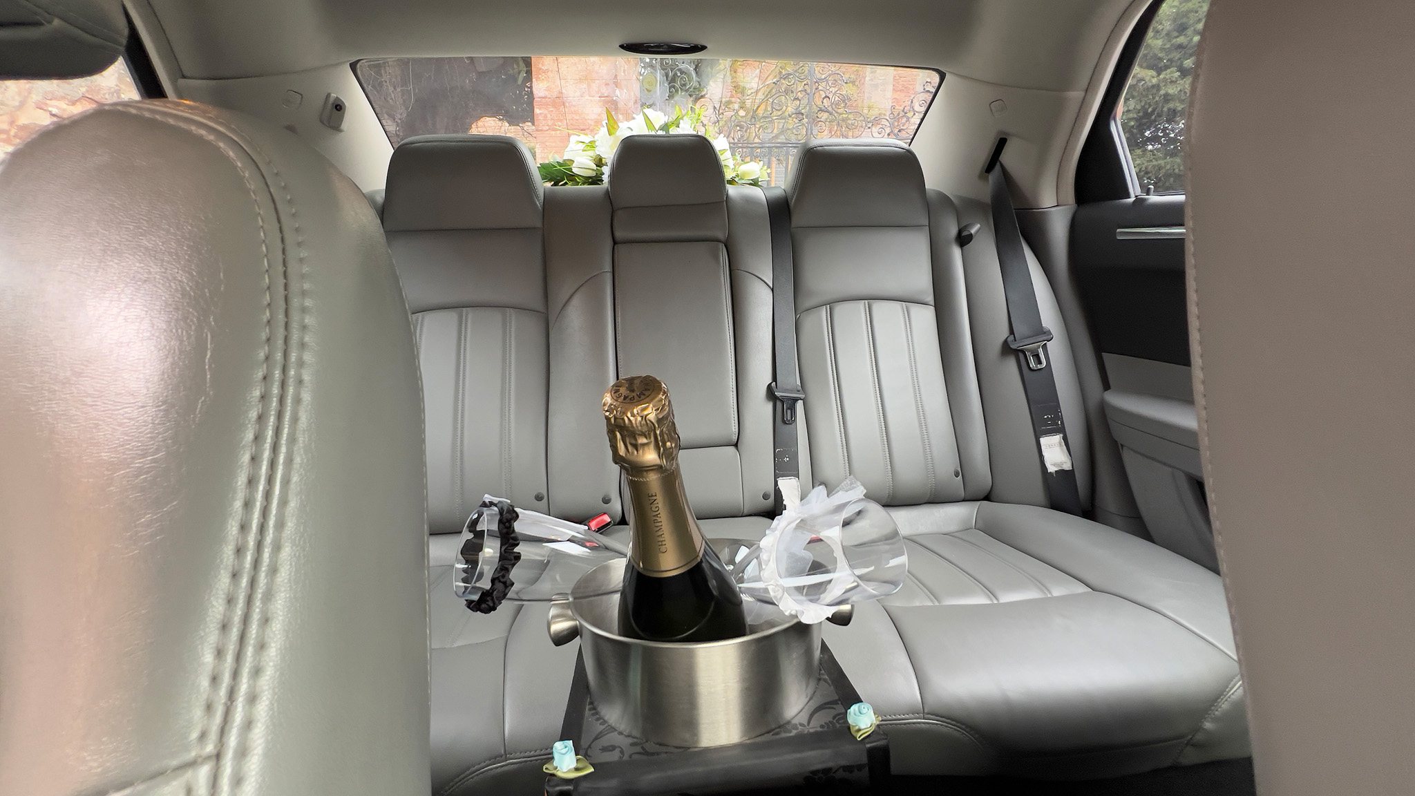 Rear interior of a Chrysler 300C wedding car showing light silver leather seats with champagne bottle and glasses placed between the rear seats.