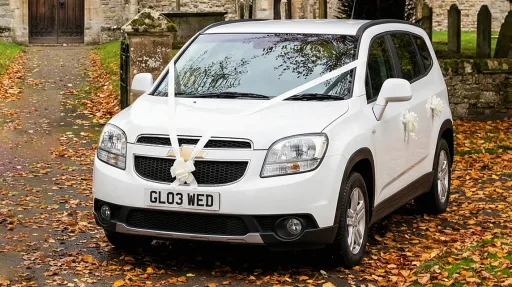 White Chevrolet Orlando wedding car decorated with white ribbons, parked on a leaf covered driveway with church buildings and trees in the background.
