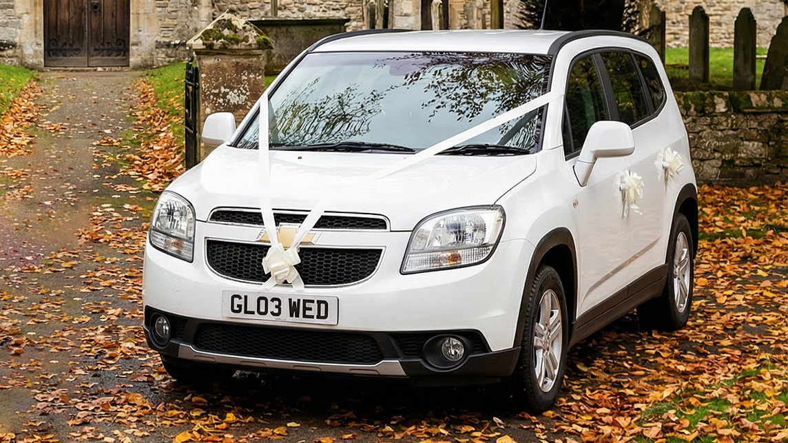 White Chevrolet Orlando wedding car decorated with white ribbons, parked on a leaf covered driveway with church buildings and trees in the background.