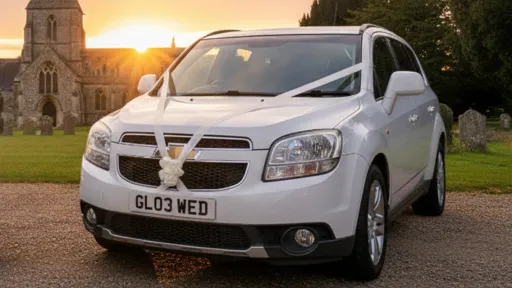 White Chevrolet Orlando wedding car with white bonnet ribbons parked on a gravel path at sunset, warm evening light illuminating the car with church and trees behind.