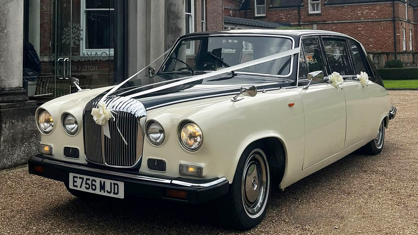 Black and ivory Daimler DS420 limousine parked on a paved driveway, front three-quarter view showing chrome grille and round headlights, finished in traditional wedding colours with clean bodywork and white ribbons and bows dressed on the vehicle