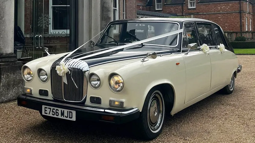 Black and ivory Daimler DS420 limousine parked on a paved driveway, front three-quarter view showing chrome grille and round headlights, finished in traditional wedding colours with clean bodywork and white ribbons and bows dressed on the vehicle