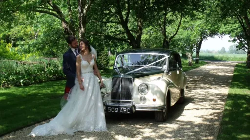 Bride and groom standing under trees beside classic Armstrong Siddeley, dappled sunlight and natural greenery.