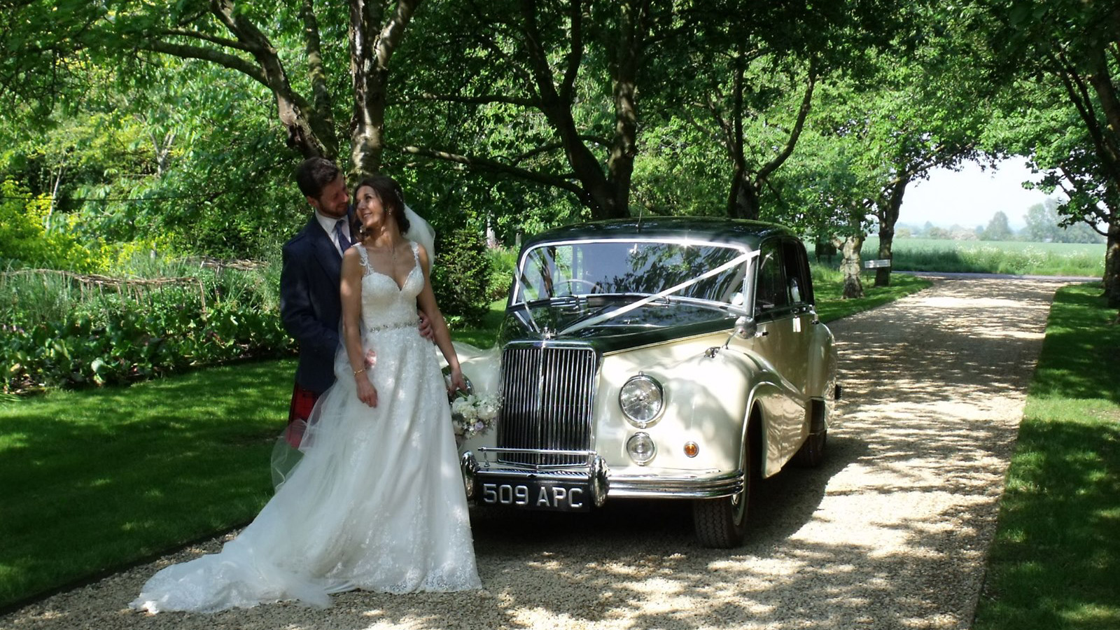 Bride and groom standing under trees beside classic Armstrong Siddeley, dappled sunlight and natural greenery.
