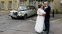 Bride in white gown and groom in dark suit standing beside a black and ivory Daimler DS420 limousine outside a stone building venue, car positioned side-on with chrome details and wedding ribbons visible on the bonnet.